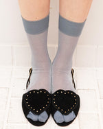 A person's legs wearing sheer light gray ankle socks with solid medium gray cuffs, paired with black open-toe heels featuring heart-shaped black velvet overlays with gold studs and light gray fabric toes, standing on a white tiled floor.