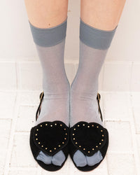 A person's legs wearing sheer light gray ankle socks with solid medium gray cuffs, paired with black open-toe heels featuring heart-shaped black velvet overlays with gold studs and light gray fabric toes, standing on a white tiled floor.
