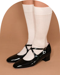 A pair of glossy black Mary Jane style low-heel shoes worn with off-white ribbed knee-high socks, shown against a solid tan background.
