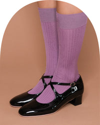 A pair of knee-high ribbed socks in a muted lavender-purple color, worn with black patent leather Mary Jane shoes with ankle straps, shown on a model's legs against a solid tan background.