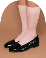A pair of light pink ribbed crew socks worn with black patent leather loafers, shown on a model's feet against a solid tan background.