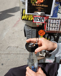 Person holding a bottle cap over a bottle with a street art-covered wall in the background.