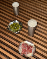 Two silver cups with a small dish of green grapes and another dish of red meat on a wooden surface.
