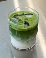 A clear glass tumbler filled with an iced layered drink, featuring a thick layer of white liquid at the bottom, a darker green middle section, and a bright green frothy top, set on a brushed metal surface.