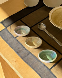 Three small handmade ceramic bowls with pastel green, blue, and beige glazes are arranged on a dark patterned placemat on a light wood table, alongside a metal spoon and other ceramic dishes.