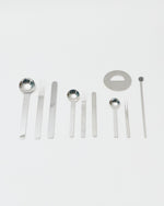 A set of stainless steel bar tools and utensils, including ladles, forks, spatulas, a circular lid or plate piece, and a long rod, arranged neatly on a light gray background.