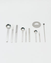 A set of stainless steel bar tools and utensils, including ladles, forks, spatulas, a circular lid or plate piece, and a long rod, arranged neatly on a light gray background.