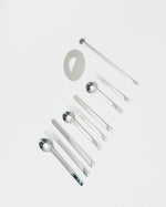 A set of polished stainless steel cutlery and tableware pieces arranged diagonally on a clean, light gray background. The set includes forks, spoons, a long stirrer, and a small oval dish.