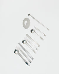 A set of polished stainless steel cutlery and tableware pieces arranged diagonally on a clean, light gray background. The set includes forks, spoons, a long stirrer, and a small oval dish.