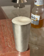 Tin beer cup on a glass surface with a bottle of beer in the background.