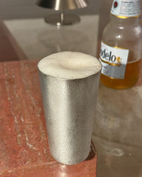 Tin beer cup on a glass surface with a bottle of beer in the background.