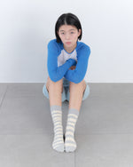 Person wearing a blue long-sleeve shirt and gray striped socks sitting on the floor against a white wall.