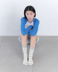 Person wearing a blue long-sleeve shirt and gray striped socks sitting on the floor against a white wall.
