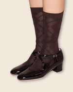 A pair of dark brown knee-high socks with a dark green argyle pattern, worn with black patent leather Mary Jane block-heel shoes, shown on a model's feet against a light beige background.