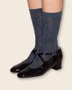 A person wearing dark blue argyle knee-high socks with black Mary Jane low-heel shoes, shown from the calves down against a solid light beige background.