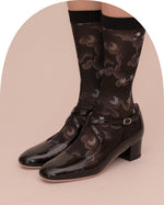 A pair of glossy black Mary Jane-style low-heel shoes with ankle straps, shown on a model wearing patterned black socks against a pale pink studio background.