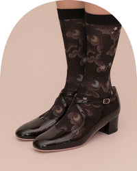 A pair of glossy black Mary Jane-style low-heel shoes with ankle straps, shown on a model wearing patterned black socks against a pale pink studio background.