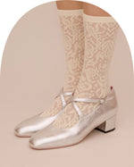A pair of metallic silver Mary Jane shoes with block heels, worn with beige patterned lace socks, shown against a solid beige background.