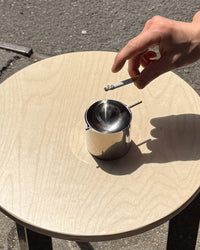 Hand holding a cigarette over a metal cup on a round wooden table outdoors.