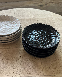 Two stacks of textured ceramic plates with scalloped edges, one black and one white, displayed on a beige stone-tiled surface.