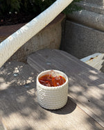 A small white textured cup filled with iced coffee and ice, placed on a weathered wooden bench outdoors.