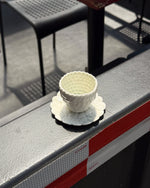 A small white textured ceramic cup with a matching saucer, placed on a gray metal railing outdoors.