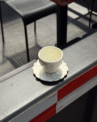 A small white textured ceramic cup with a matching saucer, placed on a gray metal railing outdoors.