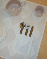 A close-up of a table setting featuring a white textured placemat with a geometric diamond pattern, a white napkin holding silver and gold-toned flatware, a white ceramic pitcher, and a white ceramic cup on a light wood table.