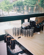 A wooden workbench with stacks of silver metal compression springs, a black marker, and office supplies, set against a large window showing a blurred street scene outside.