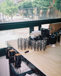 A wooden workbench with stacks of silver metal compression springs, a black marker, and office supplies, set against a large window showing a blurred street scene outside.