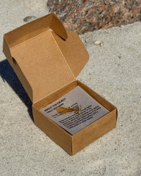 A small jewelry piece displayed on a printed card inside an open brown cardboard box, placed on a concrete surface outdoors.