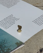 A small gold-toned metal bookmark shaped like a leaf and flower rests on an open book page outdoors on a concrete surface.