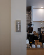 A modern wall-mounted photo frame with a brushed metal finish, shown installed on an off-white interior wall next to a kitchen or cafe counter area with coffee equipment.