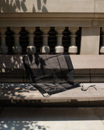 A sheer black mesh fabric accessory, possibly a scarf or small bag, draped over a light-colored wooden bench in outdoor sunlight with tree shadows.