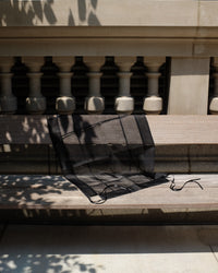 A sheer black mesh fabric accessory, possibly a scarf or small bag, draped over a light-colored wooden bench in outdoor sunlight with tree shadows.