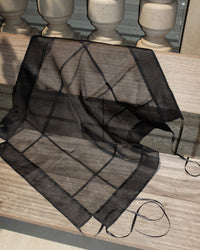 A sheer black organza scarf draped over a light-colored wooden railing, showing its fine mesh texture and grid pattern.