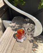 A clear glass tumbler filled with an amber-red iced drink and ice cubes, resting on a flower-shaped coaster on a weathered wooden table. The scene is outdoors with dappled sunlight and shadows, next to a white curved railing.
