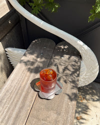 A clear glass tumbler filled with an amber-red iced drink and ice cubes, resting on a flower-shaped coaster on a weathered wooden table. The scene is outdoors with dappled sunlight and shadows, next to a white curved railing.