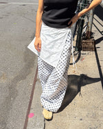 A person outdoors wearing a black sleeveless top and loose white wide-leg pants with black polka dots, standing on a sunlit sidewalk next to a bicycle.