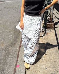 A person outdoors wearing a black sleeveless top and loose white wide-leg pants with black polka dots, standing on a sunlit sidewalk next to a bicycle.