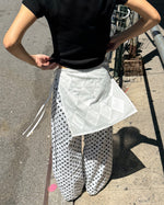 A person outdoors wearing a black short-sleeve top, white wide-leg pants with black polka dots, and a white quilted wrap or apron tied around the waist.