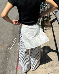 A person outdoors wearing a black short-sleeve top, white wide-leg pants with black polka dots, and a white quilted wrap or apron tied around the waist.