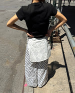 A person standing outdoors on a city sidewalk, shown from the back, wearing a black short-sleeve T-shirt, white wide-leg pants with black polka dots, and a white quilted wrap tied around the waist.
