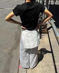 A person standing outdoors on a city sidewalk, shown from the back, wearing a black short-sleeve T-shirt, white wide-leg pants with black polka dots, and a white quilted wrap tied around the waist.
