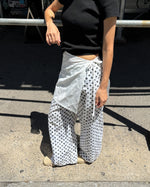 A person outdoors wearing loose white wide-leg pants with a black polka dot pattern, a black short-sleeve top, and beige shoes. The photo is taken in bright sunlight on a concrete surface.