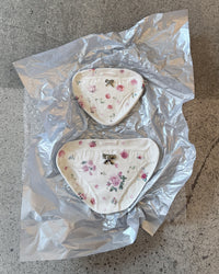 Two triangular white underwear-shaped items with pink floral prints and small metallic bow details, wrapped in crumpled silver foil on a speckled gray surface.