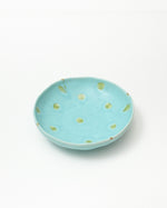 A shallow round ceramic dish in light turquoise blue with hand-painted green polka dots and small leaf motifs, photographed on a clean white background.