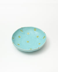 A shallow round ceramic dish in light turquoise blue with hand-painted green polka dots and small leaf motifs, photographed on a clean white background.