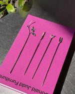 Four long black hair sticks with decorative silver metal tops, arranged on a bright pink card that reads 'Donald Judd Furniture,' photographed on a dark tabletop with a plant leaf in the corner.