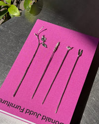 Four long black hair sticks with decorative silver metal tops, arranged on a bright pink card that reads 'Donald Judd Furniture,' photographed on a dark tabletop with a plant leaf in the corner.
